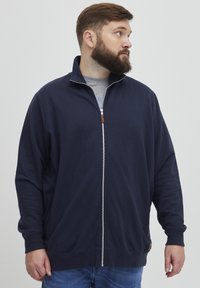 Sweat-shirt zippé marine avec un col haut, texture douce et poignets côtelés ; présente un accent en cuir près de la fermeture éclair.