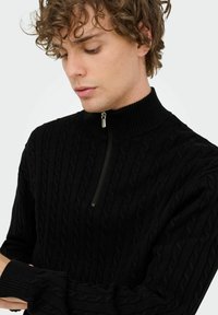 Junger Mann mit lockigem Haar, der einen schwarzen Strickpullover mit halbem Reißverschluss und geripptem Kragen trägt, und mit verschränkten Armen nach unten schaut.