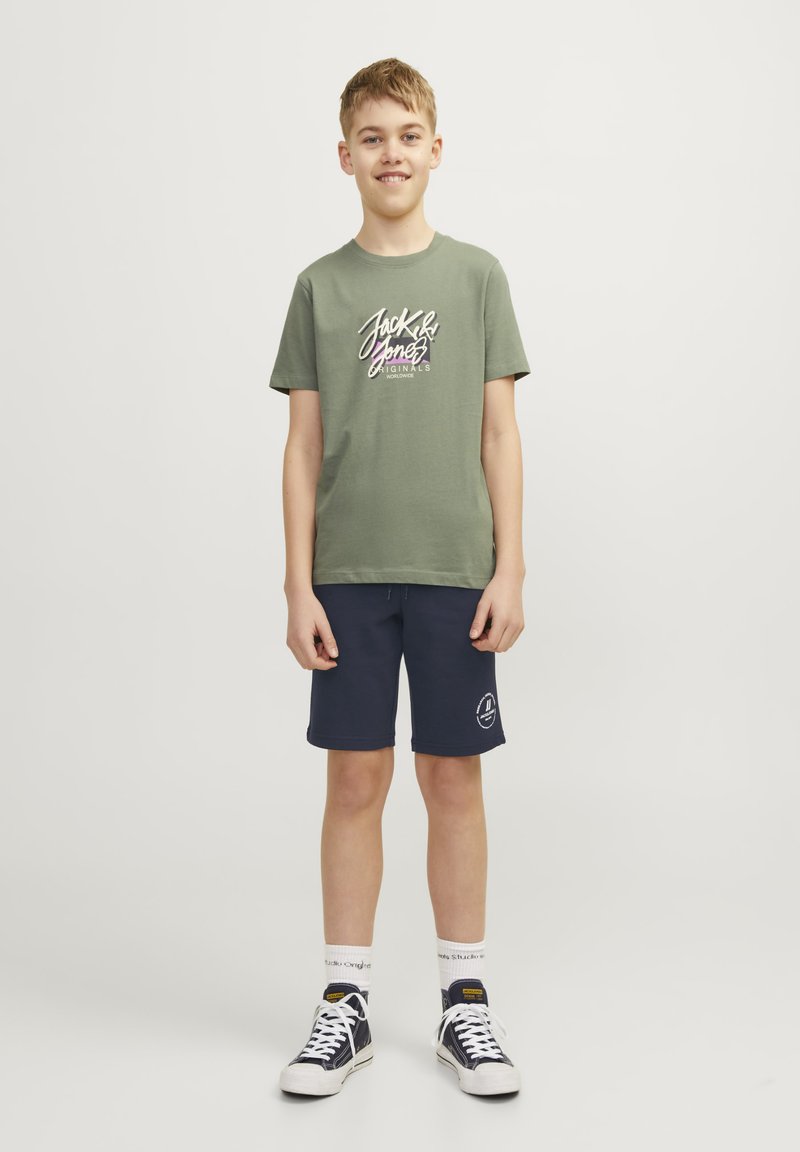Grön bomullströja med lila grafik, tillsammans med marinblå shorts och svarta sneakers. Vita strumpor med svarta detaljer. En enkel, avslappnad design.