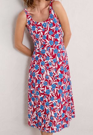 Kvinde iført ærmeløs rød kjole med blåt blomsterprint, stående mod en ensfarvet lys væg med hænderne bag ryggen.