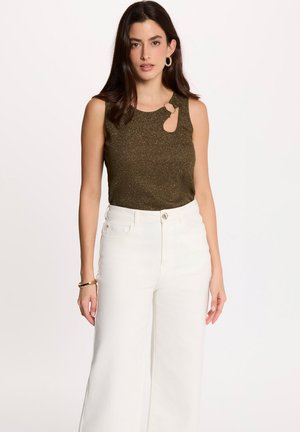 Donna con capelli scuri che indossa un top verde oliva senza maniche con dettaglio ritagliato e pantaloni bianchi a vita alta e gamba larga su sfondo bianco.
