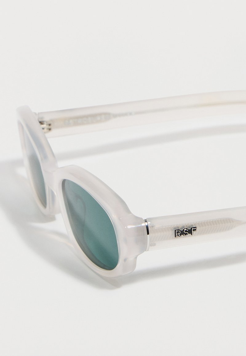 Gafas de sol con montura transparente y lentes verdes redondeadas, con las iniciales plateadas "RSF" en la patilla.