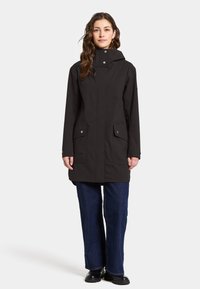 Svart vattentät jacka med huva, som har två framfickor, tryckknappar och en skräddarsydd passform, bärs över blå vida jeans.