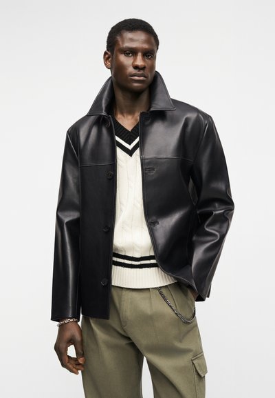 Άνδρας που φοράει μαύρο δερμάτινο μπουφάν ανοιχτό πάνω από λευκό πουλόβερ με πλέξη καλωδίου και μαύρες ρίγες, ελαι πράσινο παντελόνι cargo και αλυσίδα ως αξεσουάρ.