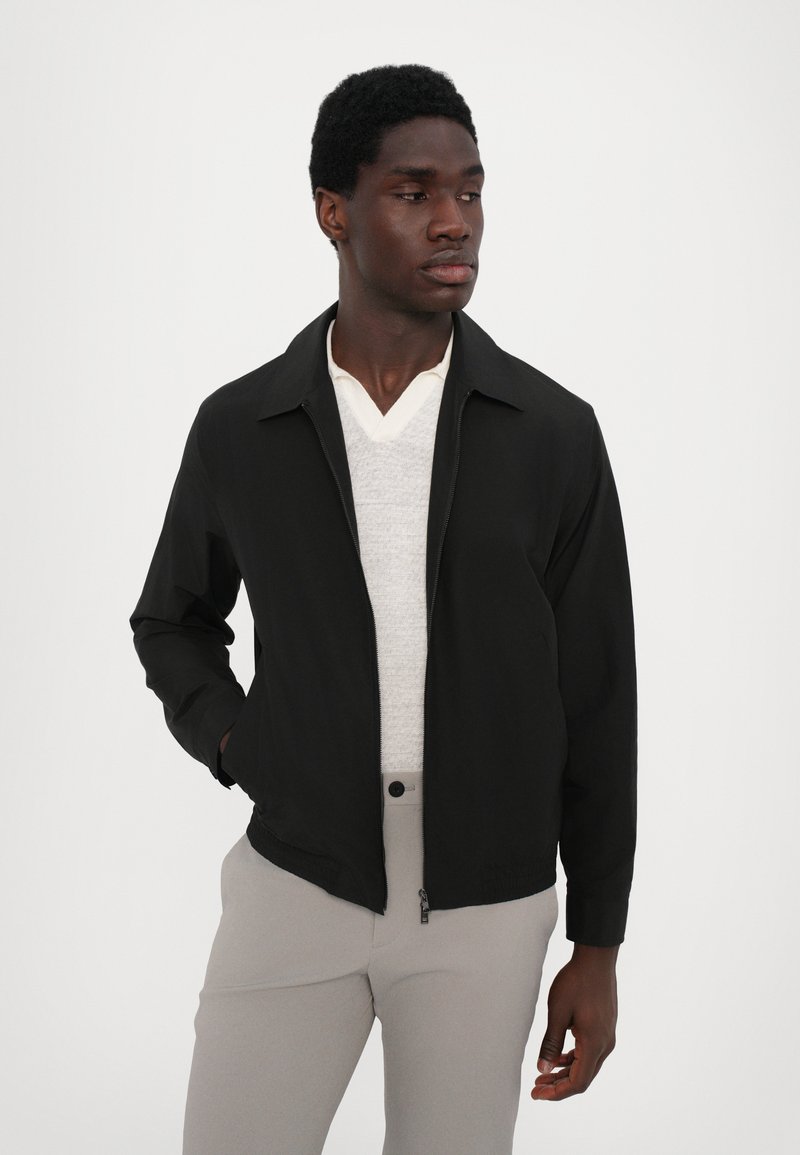 Black bomber jacket featuring a zip front, pointed collar, and long sleeves. Worn over a light gray sweater and light pants.