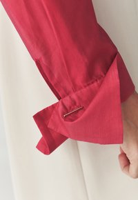 Manche en tissu texturé rouge avec un revers replié. Une épingle accentuée en or maintient le revers en place, contrastant avec un fond neutre.
