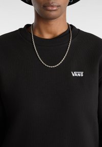 Person som bär en svart Vans-sweatshirt med ett tvinnat silverhalsband och små silverörhängen, mot en enkel bakgrund.