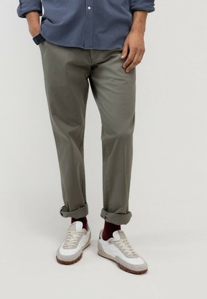 Hombre con pantalones verde oliva remangados, calcetines burdeos, zapatillas blancas y grises, y una camisa azul abotonada, de pie con las manos relajadas.