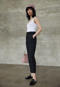 Vit långärmad bodysuit, svarta skräddarsydda byxor, glänsande svarta loafers, ljusrosa handväska och en ljusrosa bucket hat. Texturerad grå tegelvägg i bakgrunden.