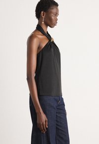 Black halter top with a knotted design at the neck and a gold accent. Smooth fabric with a relaxed fit, paired with dark blue wide-leg jeans.