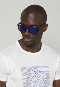 Occhiali da sole con montatura nera, lenti blu specchiate e dettagli rossi, abbinati a una camicia bianca con un motivo a righe blu.