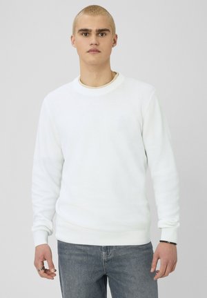 Jeune homme aux cheveux blonds courts, portant un pull blanc texturé et un jean bleu, debout devant un fond neutre.