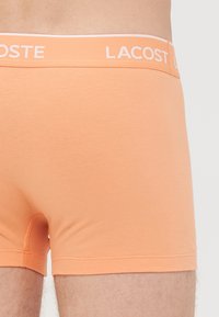 Close-up van een persoon die perzikkleurige Lacoste boxershort met een gemarkeerde elastische tailleband draagt.