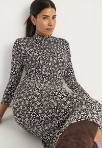 Langsleeve, aansluitende jurk in zwart met een bloemenpatroon in crèmekleurige en bruine tinten. Hoge halslijn en zachte, rekbare stof.