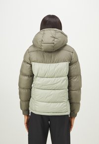 Pufota jaka olīvzaļā un gaiši zaļā krāsā, ar kapuci, quilted dizainu un elastīgām piedurknēm, ar gludu tekstūru.
