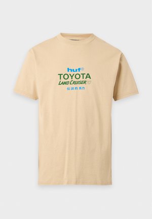 Un mannequin aux cheveux tressés est de dos, montrant un T-shirt HUF beige orné de motifs Toyota Land Cruiser et un pantalon assorti.