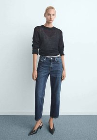 Pull en maille noir à manches longues, associé à un jean droit bleu. Des escarpins noirs à bout pointu complètent la tenue. Fond clair.