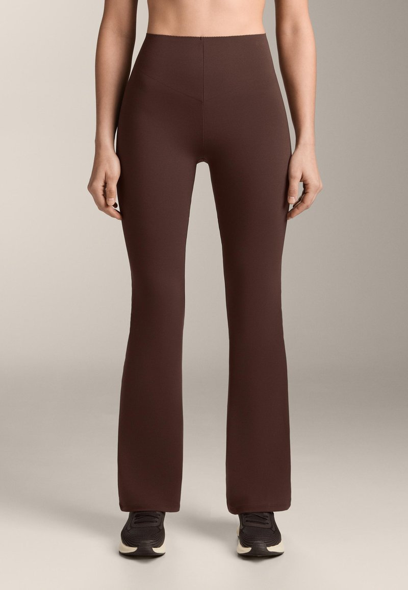Personne portant un pantalon ajusté taille haute, marron foncé avec une légère évasure, associé à des baskets noires et blanches, debout devant un fond neutre.