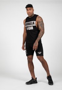 Zwarte mouwloze tanktop met de tekst "GORILLA WEAR", gecombineerd met zwarte shorts met logo's. Het model draagt zwarte sportschoenen.