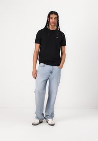 T-shirt en coton noir avec un petit logo, associée à un jean décontracté bleu clair et à des baskets blanches avec des accents noirs. Tenue simple et décontractée.