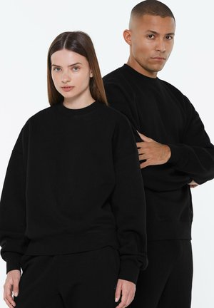 A man and woman stand side by side, both wearing plain black sweatshirts and black pants against a white background.