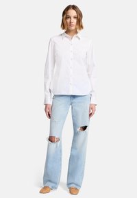 Vit skjorta med långa ärmar parat med ljusblå slitna utsvängda jeans med stora knäskåror och ljusbruna slip-on skor.