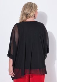 Czarna bluzka z szyfonu z przezroczystymi rękawami, luźnym krojem i linią dołu sięgającą poniżej bioder. Połączona z czerwonymi spodniami o teksturowanej powierzchni.