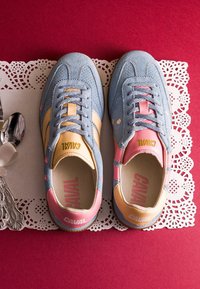 Paire de baskets en daim bleu avec des accents roses et jaunes, marquage « CAVAL » sur la languette et les talons, posées sur un napperon en dentelle blanche sur fond rouge.
