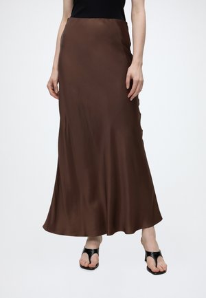Femme portant une jupe longue en satin marron avec un haut noir et des sandales à talons ouvertes noires sur un fond blanc uni.