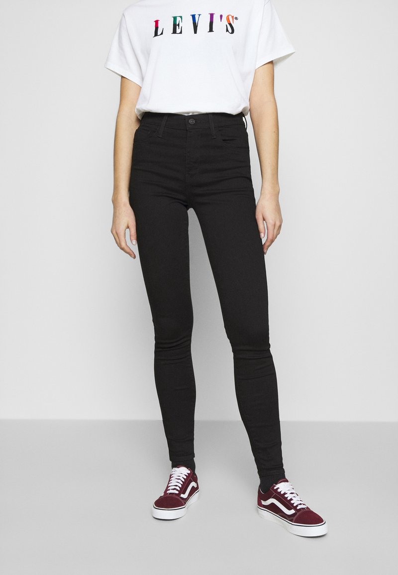 Jeans skinny de cintura alta negros combinados con una camiseta blanca que presenta un colorido logo de "Levi's", complementados por zapatillas burdeos.