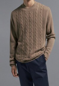 Beige Kabelstrickpullover mit Rundhalsausschnitt, strukturiertem Muster und gerippten Bündchen, kombiniert mit navyfarbigen Hosen. Gemütliches und strukturiertes Design.