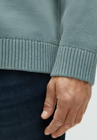 Hellblauer Strickpullover mit gerippten Bündchen. Die Textur ist glatt und die Passform wirkt entspannt. Die Hand liegt nahe an der dunkelblauen Jeans.