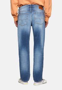 Jeans de mezclilla azul con un corte relajado, ligero desvanecimiento y dos bolsillos traseros. La cintura presenta una etiqueta con el logo marrón de Diesel. Se llevan zapatillas blancas.