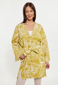 Keltainen kimono-tyylinen takki, jossa on valkoisia lehtikuvioita, pitkät hihat ja vyötärölle sidottu vyö. Kevyt kangas ja rento istuvuus.