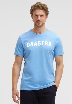Man with light brown curly hair wearing a light blue Gaastra t-shirt and dark pants, standing with one hand in pocket against a plain background.