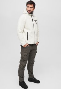Giacca bianca in pile con zip e cappuccio, dettagli neri; abbinata a pantaloni cargo verde oliva con molte tasche. Stivali neri.