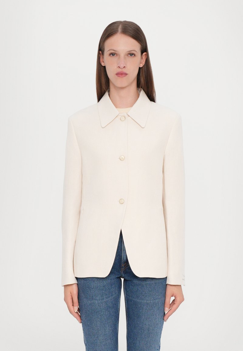 Cream-colored jacket with a collar, front buttons, and side pockets. Smooth fabric, structured shape, and tailored fit. Worn over blue jeans.