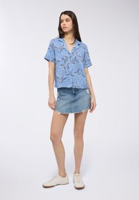 Camicia a maniche corte blu con fantasia floreale, abbinata a una gonna di jeans sfilacciata. Il modello indossa sneakers bianche, con una vestibilità rilassata e dettagli ben definiti.