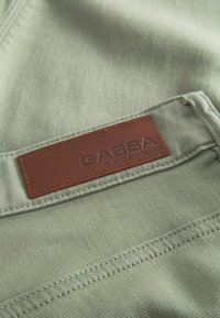 Olivovo zelená látka s hladkou textúrou, na ktorej je hnedá kožená nášivka s vyrazeným textom "GABBA" a "RELATABLE SINCE 1983."