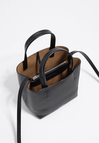 Zwarte leren handtas met dubbele handgrepen, een binnenvak met ritssluiting en een binnenzijde van bruin suede, opengevouwen op een witte ondergrond.