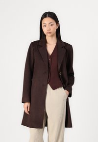 Manteau long marron avec un design structuré, revers crantés, deux boutons et une texture douce, porté sur un gilet bourgogne et un pantalon clair.