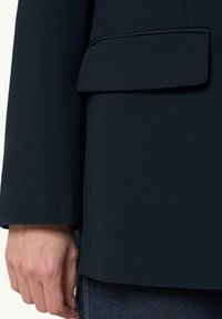 Marineblauwe op maat gemaakte blazer van een gestructureerde stof, met een borstzak en een strakke, gestructureerde uitstraling. Hand rust op de rand van de blazer.