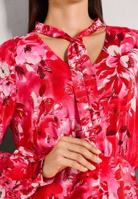 Rote Bluse mit Blumenmuster und einer Schleife am Kragen, mit großen rosa und weißen Blütenmustern. Lange Ärmel mit geknöpften Manschetten, glatte Stoffstruktur.