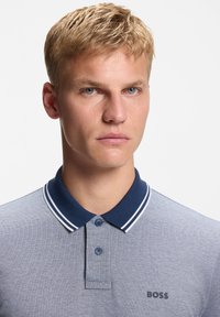 Νέος άνδρας με κοντά ξανθά μαλλιά, φοράει γαλάζιο πόλο μπλουζάκι με σκούρο μπλε γιακά και το λογότυπο "BOSS" στο στήθος.