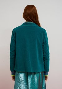 Petrolfarvet ribjakke med reverskrave og lange ærmer, med ribstrikkede manchetter i kontrastfarver, båret over et skinnende sølvfarvet skørt.