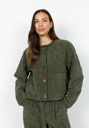Woman with long brown hair wears olive green corduroy jacket and matching pants, standing with hands in pockets against white background.