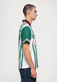 Green and white striped short-sleeve polo shirt with a black collar, red accents, and a logo on the front. Fabric appears smooth and lightweight.