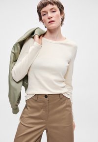 Απαλή κρεμ μπλούζα με μακριά μανίκια, φτιαγμένη από μαλακό ύφασμα, σε συνδυασμό με καφέ κομμένα παντελόνια και μια ελαιοπράσινη μπομπέρ τζάκετ, κρατημένη πάνω στον ώμο.