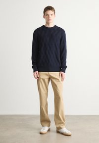 Maglione lavorato a maglia navy con un motivo a diamante testurizzato, abbinato a pantaloni beige chiaro e sneakers bianche, in piedi su uno sfondo neutro.