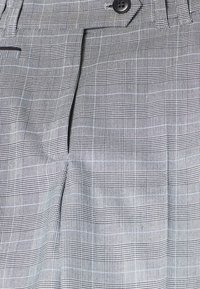 Gros plan sur un pantalon à carreaux gris avec un bouton et une braguette zippée, présentant un motif à carreaux subtil composé de fines lignes blanches et noires.